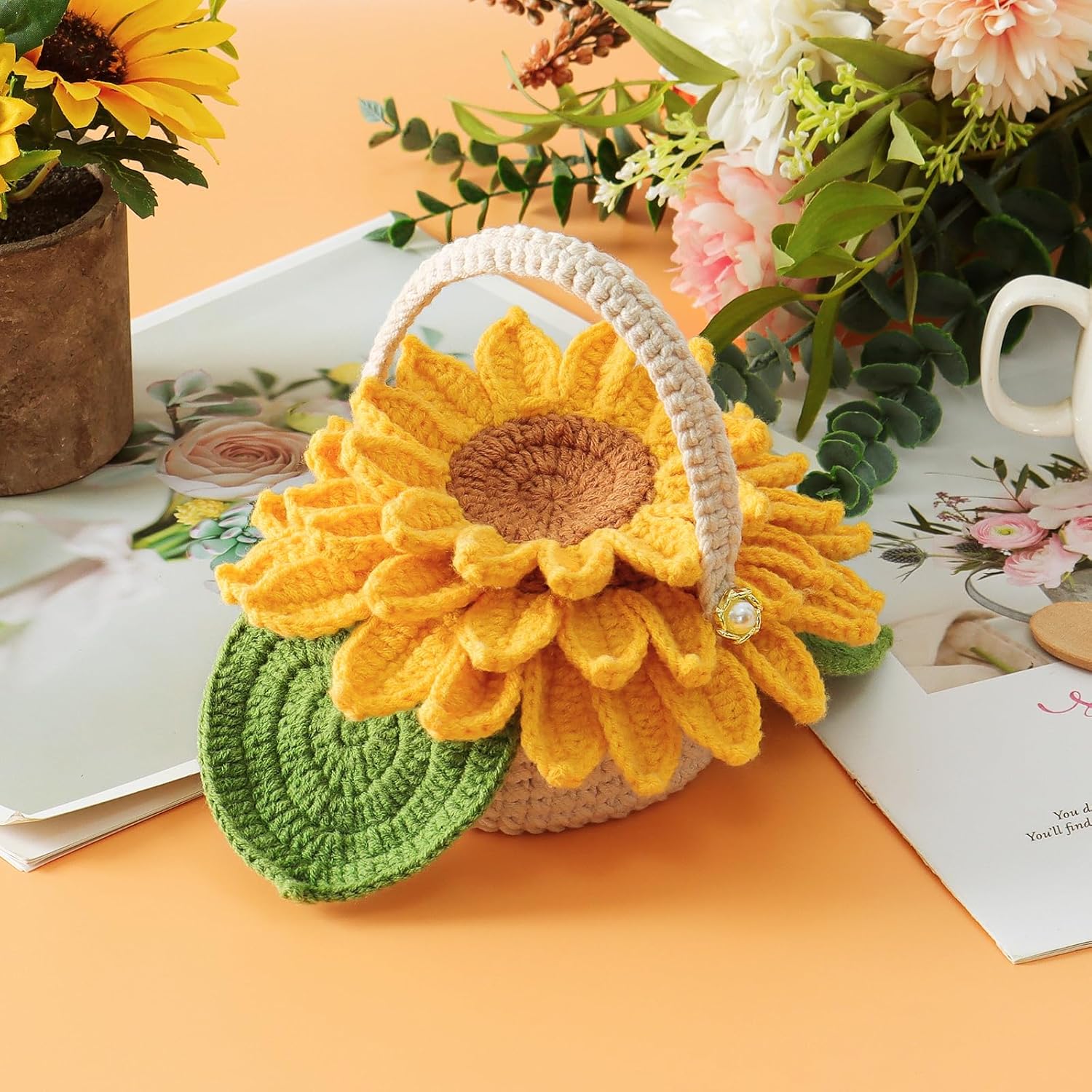 The Sunflower Pot Crochet Kit with 5pcs Coasters features a crocheted sunflower basket with green leaves, displayed on a table alongside handmade sunflower coasters, flowers, a mug, and an open magazine.