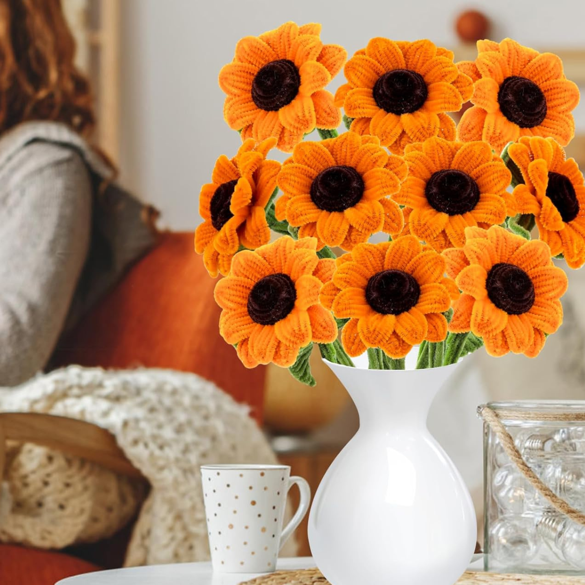 A Sunflower Crochet Kit vase sits on a table beside a white mug with gold dots and a glass jar, with someone seated in the background.