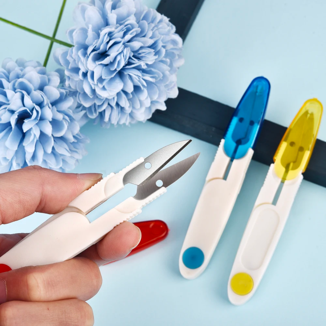A hand holding Yarn Thread Snips with a protective cover and a colorful selection of Yarn Thread Snips and a blue artificial flower in the background.