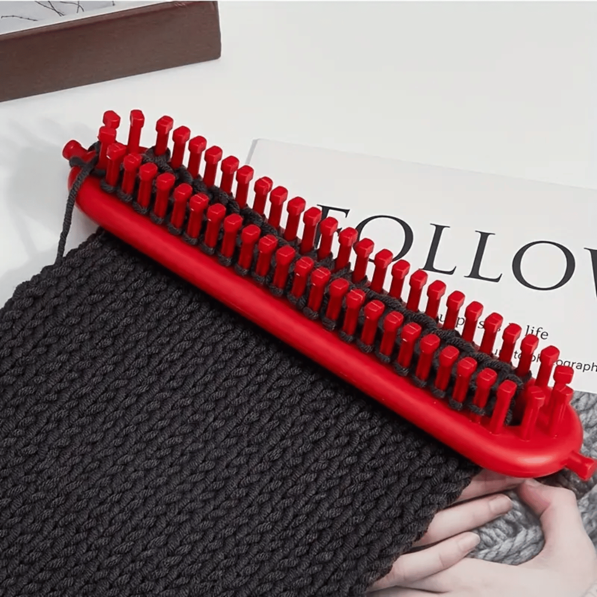 The Knitting Loom Set, beginner-friendly and rectangular in red, holds dark gray yarn with fast knitting in progress on a magazine showing "FOLLOW." Ideal for quick and easy knitting projects.