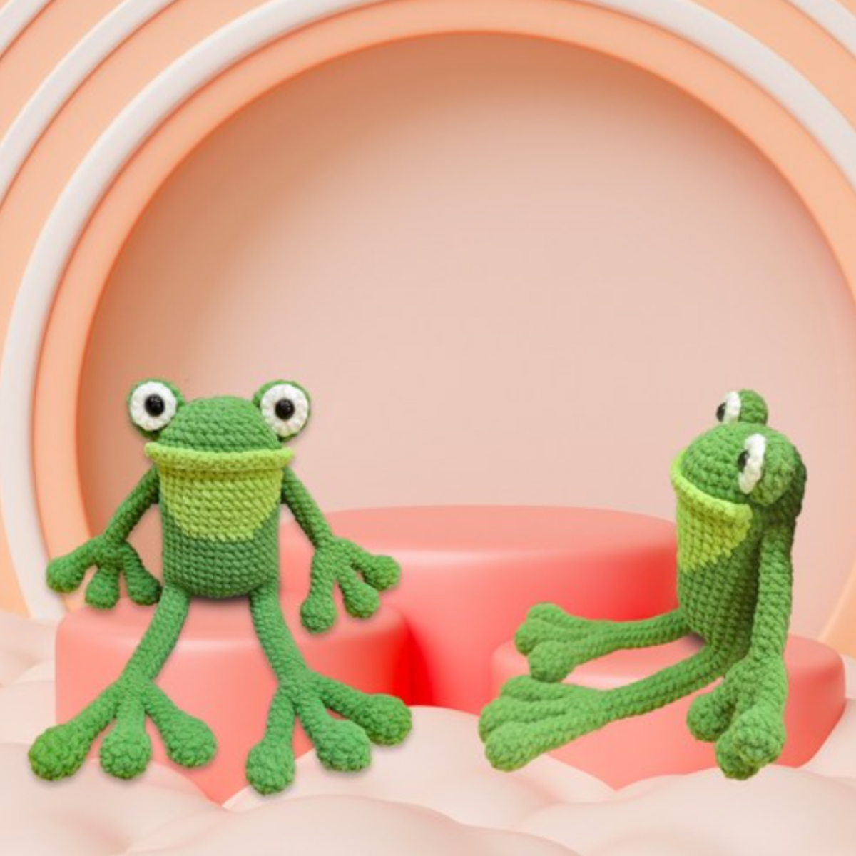 Two amigurumi frogs made with the Crochet Frog Kit are displayed on a pink abstract set featuring arches and rounded platforms in the background.