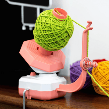 A pink and white Electric Yarn Winder holds a green yarn ball, with yellow and purple yarn balls in the background on a wooden surface.
