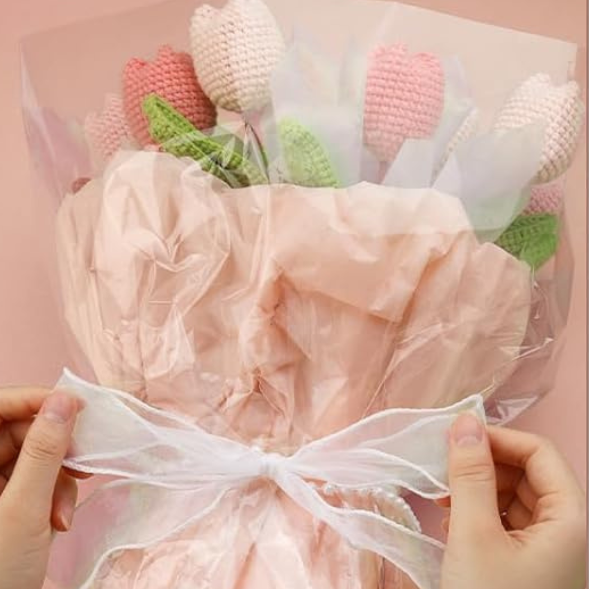 Hands tying a ribbon around a bouquet of pink, white, and green knitted flowers wrapped in pink and clear plastic—an adorable Crochet Tulip Bouquet Kit for an everlasting tulip crochet gift.