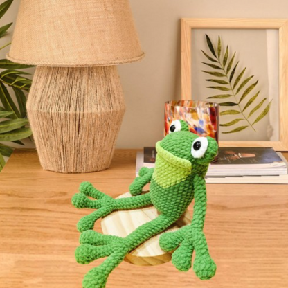 A Crochet Frog Kit plush sits on a wooden surface near a lamp, a leaf-patterned glass, and a framed leaf print—perfect inspiration for your next beginner crochet project.