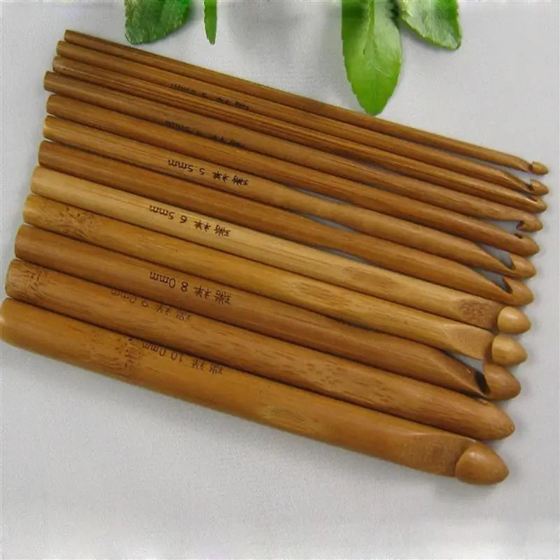 A set of Bamboo Crochet Hooks 12 Pcs of varying sizes arranged in a straight row on a white surface, with green leaves in the background. Perfect for sustainable crafting, these eco-friendly tools are both functional and stylish.