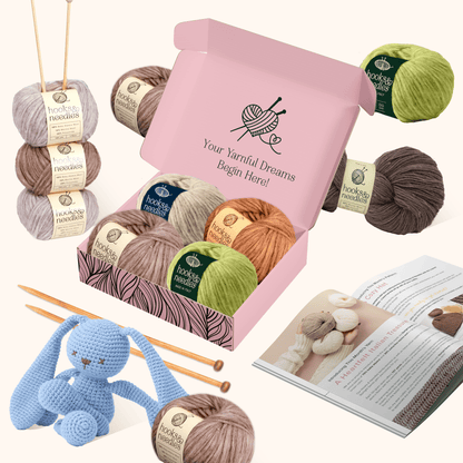 A 6-Month-Prepaid Hooks & Needles Subscription Box #14 featuring yarn balls, crochet hooks, a pattern book, and a blue crocheted rabbit, arranged with an open pink box on a light background.