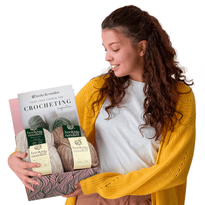 Woman in a yellow cardigan smiling at a Hooks & Needles Subscription Box #30, standing against a white and green background.
