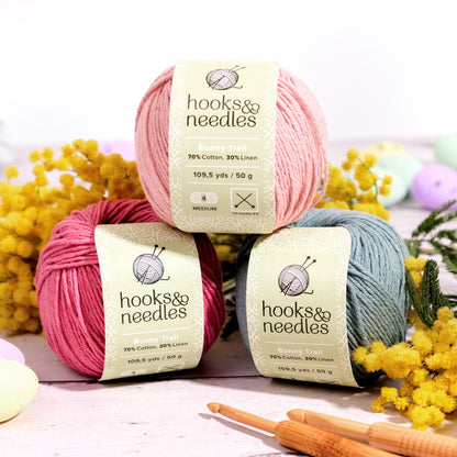 Three balls of Limited Edition Easter Special Collection (bundle) with a pair of knitting needles and decorative yellow flowers on a wooden surface.