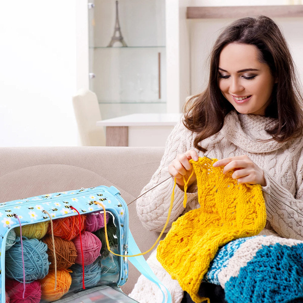 A woman with long dark hair is sitting on a couch, knitting a yellow piece. Various colorful yarn balls are stored in a spacious Yarn Bag - Storage Organizer next to her. A shelf and Eiffel Tower figurine are in the background.