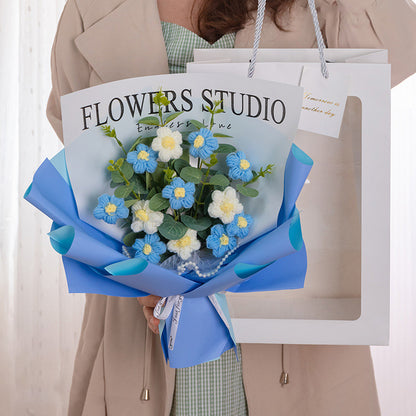 A person holds a Sunny Crochet Daisy Bouquet: Forever in Bloom, artfully arranged with blue and white flowers wrapped in blue paper, alongside a shopping bag labeled "FLOWERS STUDIO.