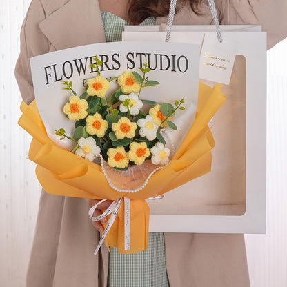 A person holding the "Sunny Crochet Daisy Bouquet: Forever in Bloom," an exquisite arrangement of yellow and white flowers wrapped in orange paper, alongside a shopping bag labeled "Flowers Studio," exudes the charm of everlasting beauty. Each handcrafted floral detail captures the essence of nature’s grace.