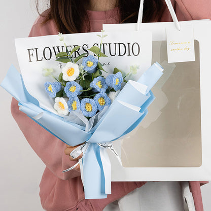 A person holds an elegant gift bag containing the "Crochet Lilies of the Valley Bouquet: Handcrafted Grace," featuring a mix of meticulously crafted blue and white flowers delicately accented with crochet lilies. A note peeks out, reading "Flowers Studio.