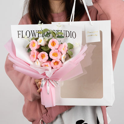 A person holds an elegant gift bag emblazoned with "FLOWER STUDIO," containing a Crochet Lilies of the Valley Bouquet: Handcrafted Grace, featuring pink, knitted flowers wrapped in pink paper.