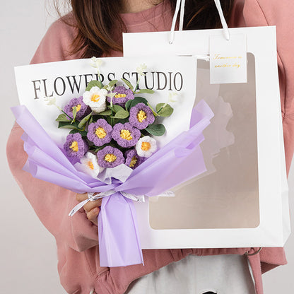 A person holds the Crochet Lilies of the Valley Bouquet: Handcrafted Grace, an elegant arrangement of purple and white flowers wrapped in lilac paper, accompanied by a stylish white paper bag with a clear window.