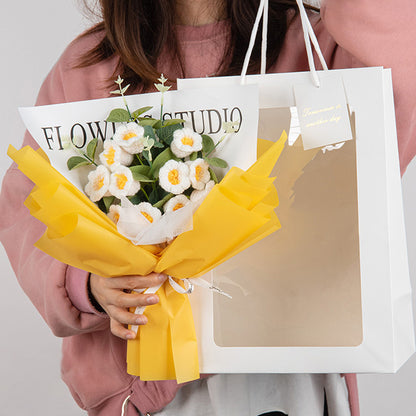 A person holding an elegant gift: the "Crochet Lilies of the Valley Bouquet: Handcrafted Grace," featuring white crocheted flowers with yellow accents, wrapped in yellow paper, and accompanied by a white gift bag labeled "Flower Studio.