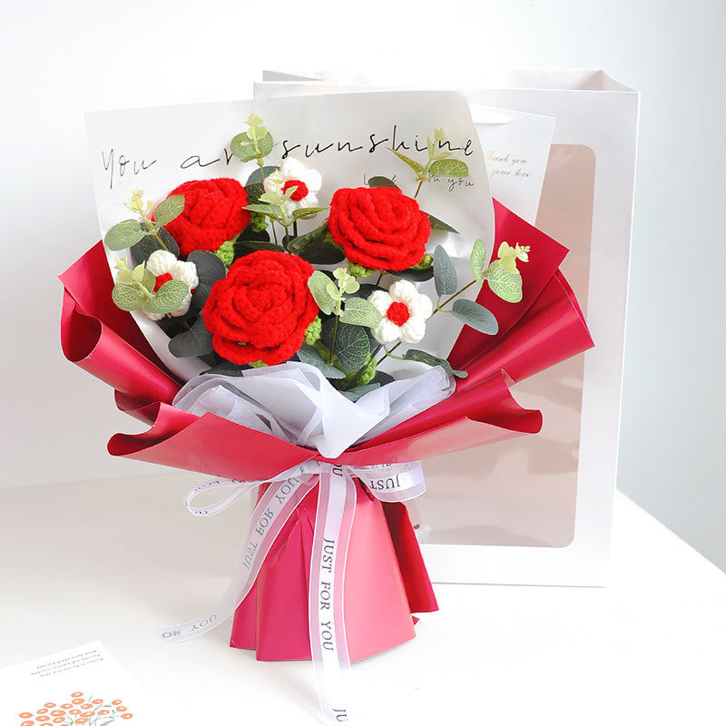 A bouquet of handcrafted roses, white flowers, and green leaves wrapped in red and white paper with a ribbon that reads "Just for you." This display of timeless elegance is complemented by a partially visible card behind the Crochet Rose Bouquet - Timeless Blooms.
