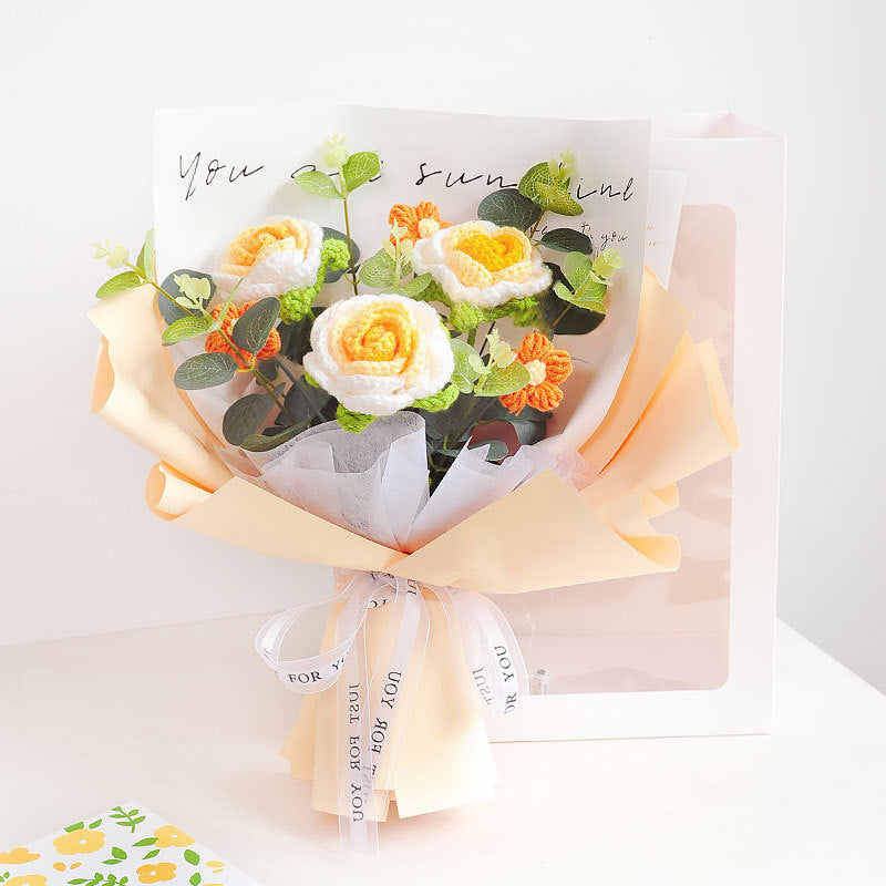 The Crochet Rose Bouquet - Timeless Blooms (Bundle51), featuring handcrafted crocheted yellow and white flowers with delicate green leaves, elegantly wrapped in peach-colored paper and adorned with a ribbon, showcases timeless elegance against a white background.