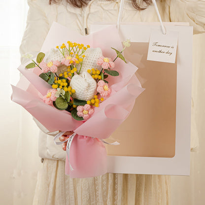 A person holds a Crochet Tulip Bouquet: Springtime in Every Stitch, featuring handcrafted tulips of pink and yellow hues, wrapped in pink paper, beside a white gift bag with a tag that reads "Tomorrow is another day.