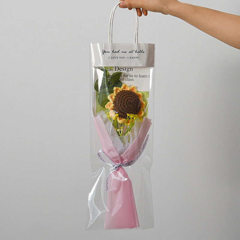 A hand holds a clear gift bag containing a sunflower with green leaves wrapped in pink paper, alongside the Small Bouquet of Crochet Flowers.