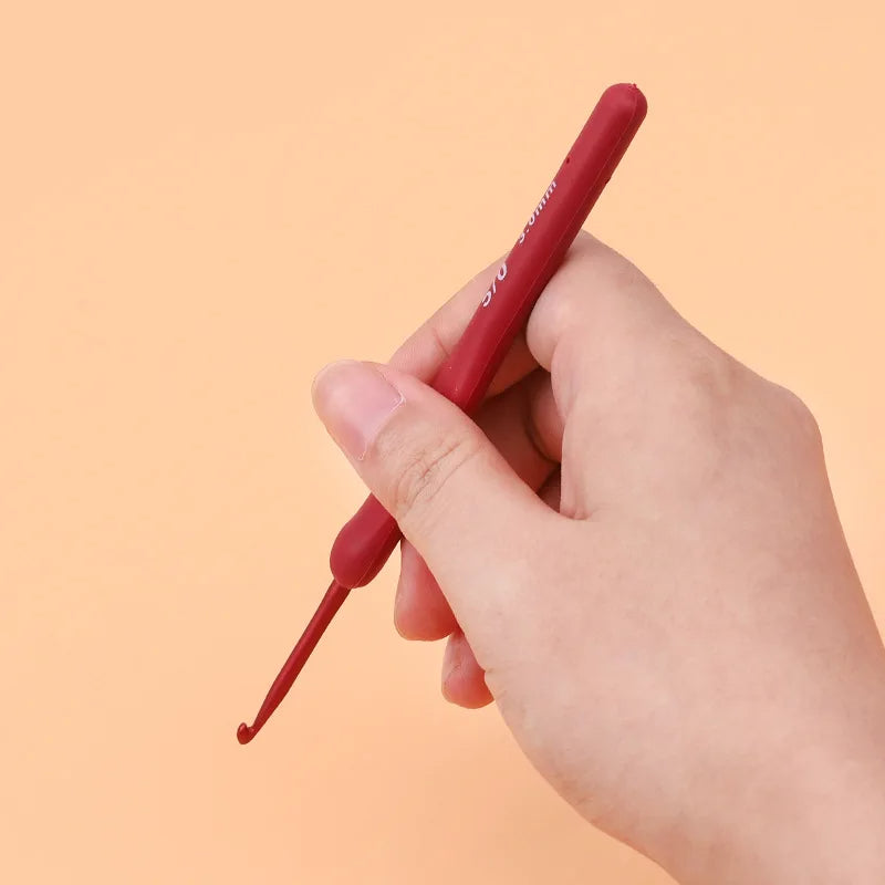 A hand holds a red crochet hook from the Ergonomic Crochet Hook Set 9 Pcs with Soft Grip Handles against a peach-colored background, ensuring comfortable crocheting throughout any project.