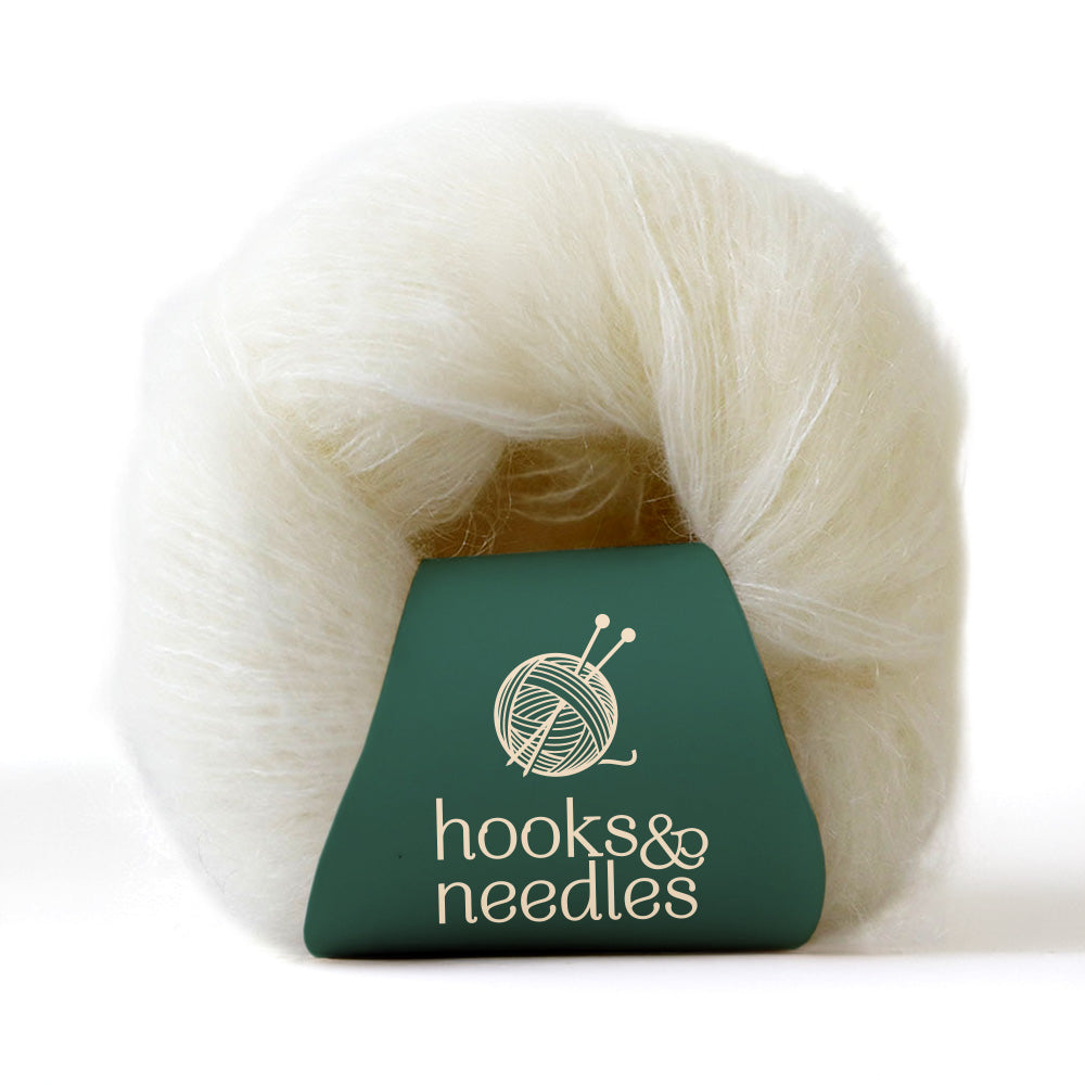 A skein of Fusion Finesse Yarn 20g in white lace-weight, adorned with a green "hooks & needles" label and a knitting logo, provides the delicate touch and elegance of mohair.