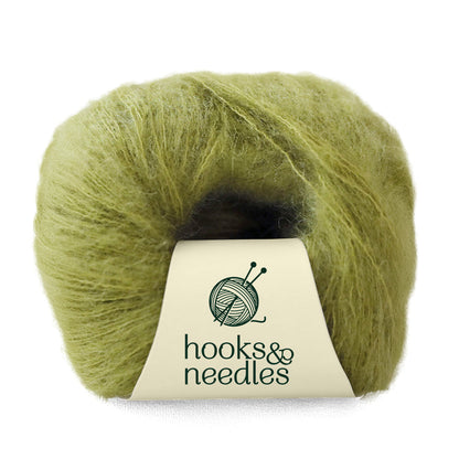 A skein of Fusion Finesse Yarn 20g, offering soft, fuzzy green fibers ideal for lace-weight projects, is labeled with "hooks & needles" and blends luxurious materials such as mohair and merino wool.