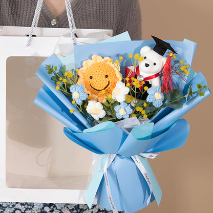 A person holds the Doctor Bear Graduation Season Crochet Bouquet, featuring a blue wrapping, a smiling sun plush, a white bear with a graduation cap, and small flowers. This charming bouquet perfectly captures the essence of the special occasion.