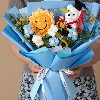 A person holds a Doctor Bear Graduation Season Crochet Bouquet, which includes a sun, a doctor bear in a graduation cap, and blue and white flowers, all wrapped neatly in blue paper.