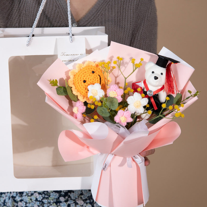 A person holds a gift bag and a crochet bouquet, wrapped in pink, showcasing the "Doctor Bear Graduation Season Crochet Bouquet," which includes a crocheted lion and small flowers, with the Doctor Bear wearing a graduation cap and gown.