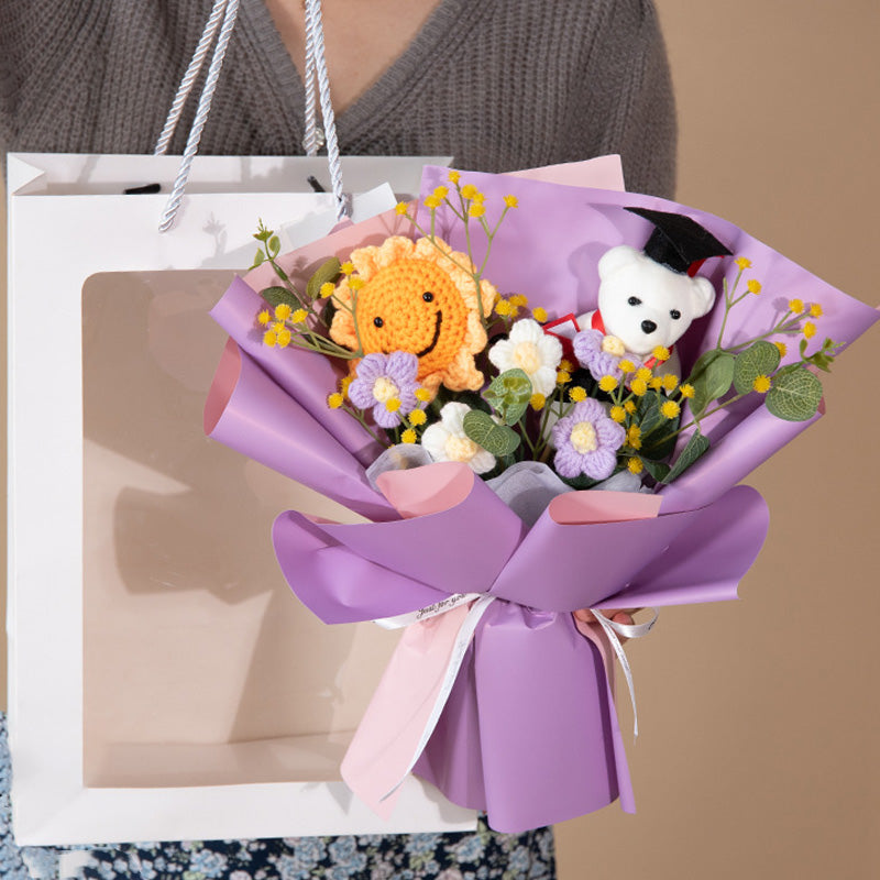 A person delightedly holds a gift bag with the Doctor Bear Graduation Season Crochet Bouquet, featuring a vibrant array of purple and yellow flowers and a crocheted lion.