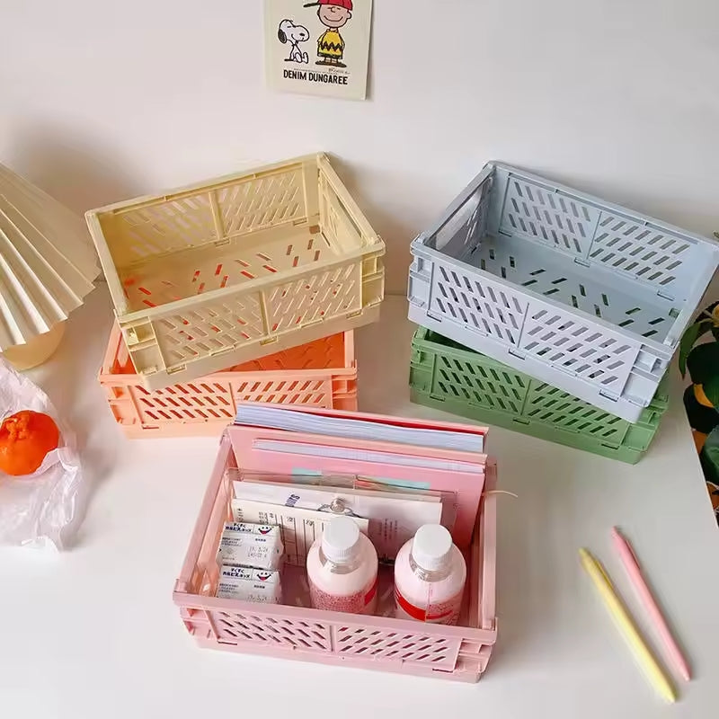 A set of four vibrant, stackable plastic storage crates are displayed on a table, with one crate neatly holding various items. The Collapsible Storage Crate (10" x 7") for Knitting / Crocheting Supplies is ideal for organizing crocheting accessories. Nearby are a lamp, some fruit, pens, and a cartoon photo.