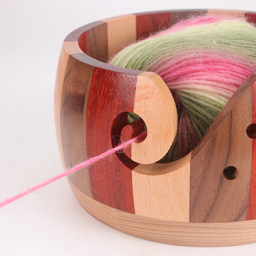 A ball of multicolored yarn in a Handmade Wooden Yarn Bowl with a thread passing through a swirl-shaped cutout.