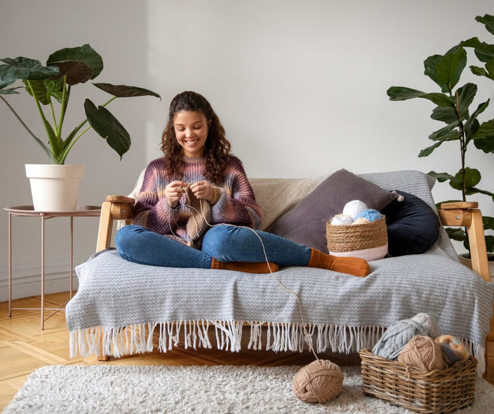 Woman sitting on a couch knitting with a basket of yarn beside her.