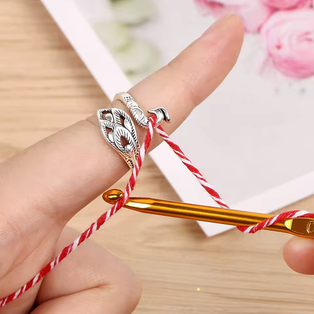 A hand with silver rings uses the Yarn Tension Control Ring to hold red and white yarn and a gold crochet hook, starting a crochet project on a wooden surface.