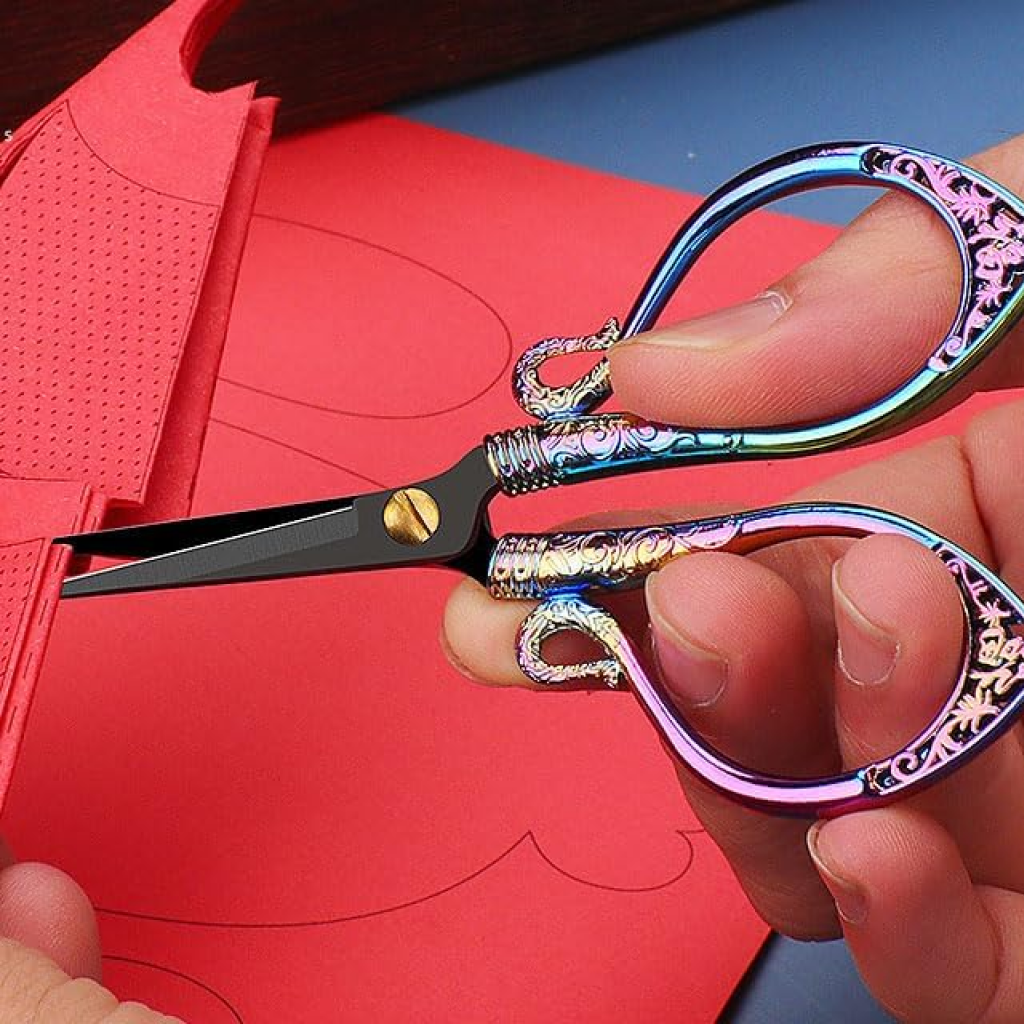A hand uses Decorative Vintage Embroidery Scissors with ornate, multicolored handles to cut patterned red paper—ideal for precision crafting.