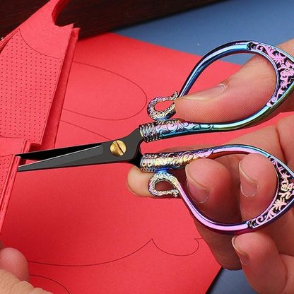 A hand uses Decorative Vintage Embroidery Scissors with ornate, multicolored handles to cut patterned red paper—ideal for precision crafting.