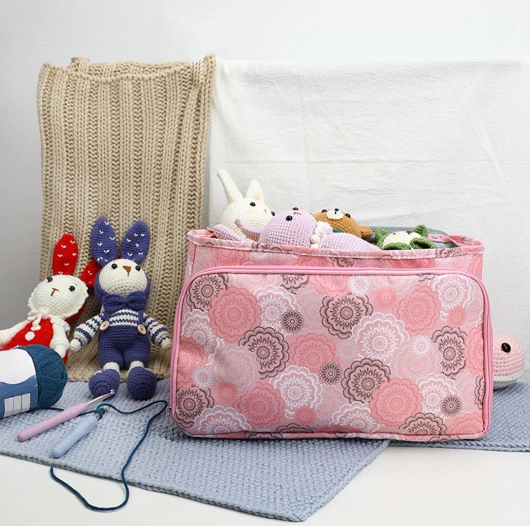 A Multi-Purpose Craft Bag in a pink pattern, filled with stuffed animals, sits on a gray mat, surrounded by knit blankets, crochet tools, and plush toys.