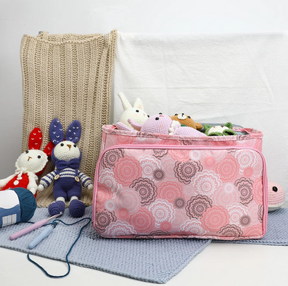 A Multi-Purpose Craft Bag in a pink pattern, filled with stuffed animals, sits on a gray mat, surrounded by knit blankets, crochet tools, and plush toys.