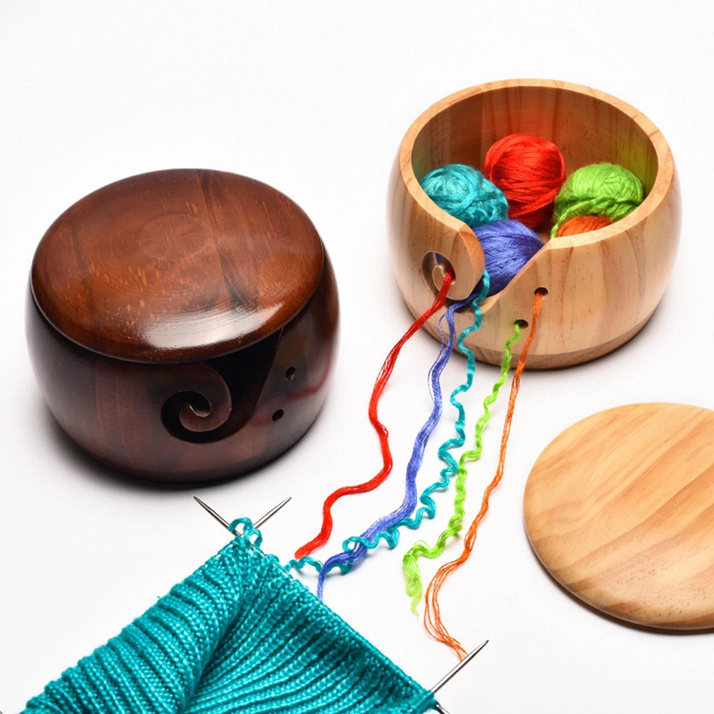 A Wooden Yarn Bowl With Lid holds colorful yarn balls with unwinding strands beside knitting needles working on turquoise fabric, all displayed on a white background.