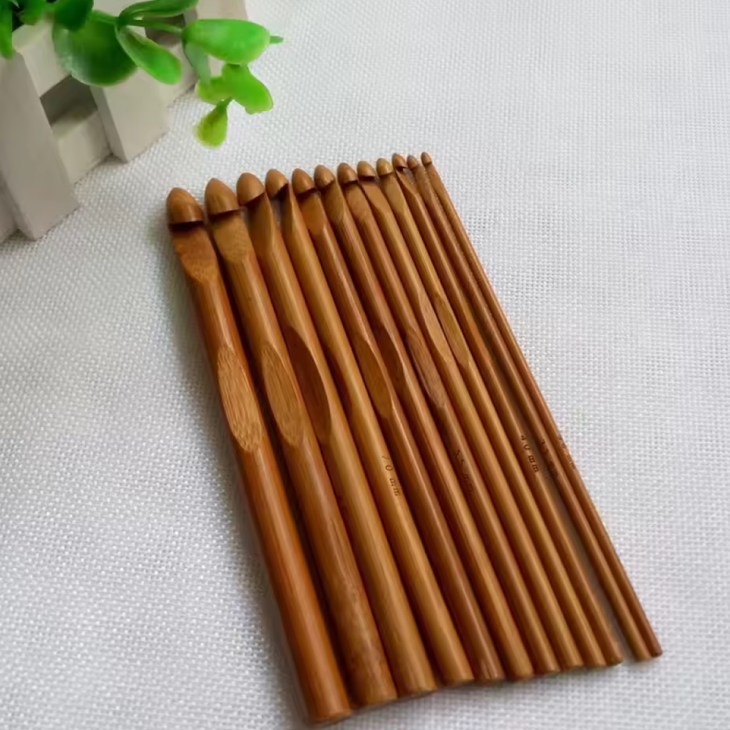 A set of twelve eco-friendly Bamboo Crochet Hooks 12 Pcs in assorted sizes is neatly arranged on a light textured surface beside a green leafy plant.