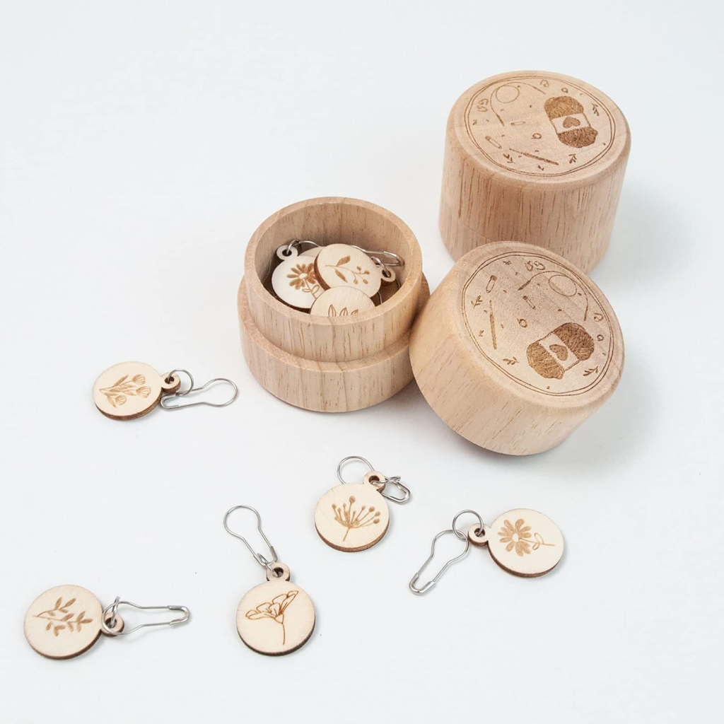 The Wooden Flower Stitch Markers with Storage Box 8Pcs set includes eight durable wooden stitch markers, each with a unique botanical design, stored in round engraved-lid boxes. Some markers are displayed outside the containers.