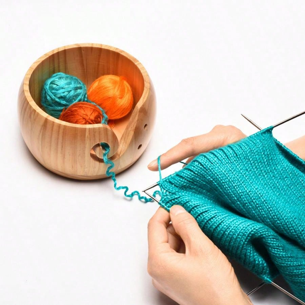 Hands knitting with teal yarn and metal needles. A Wooden Yarn Bowl With Lid, featuring a high gloss finish, holds blue, orange, and gold yarn balls on a white surface.