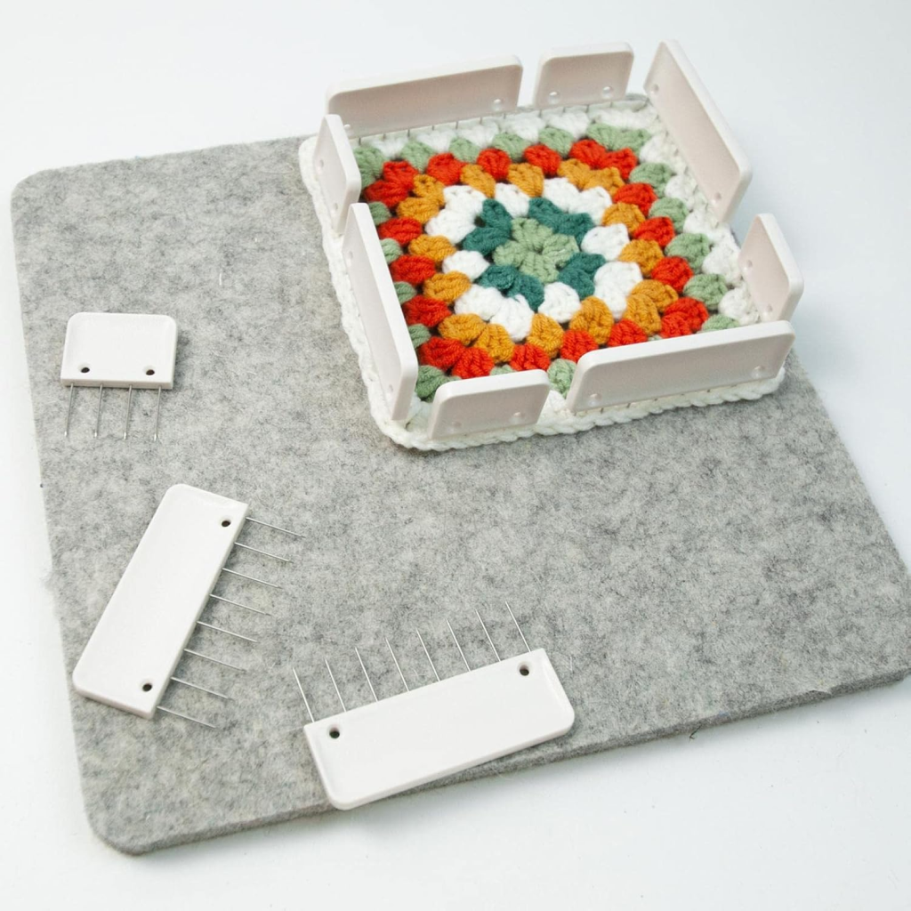 A multicolored crocheted granny square is held by a white blocking frame from the Knit Blocking Comb Set on a gray felt mat, with two frame pieces, pins, and a knit blocking comb nearby for flexible project use.