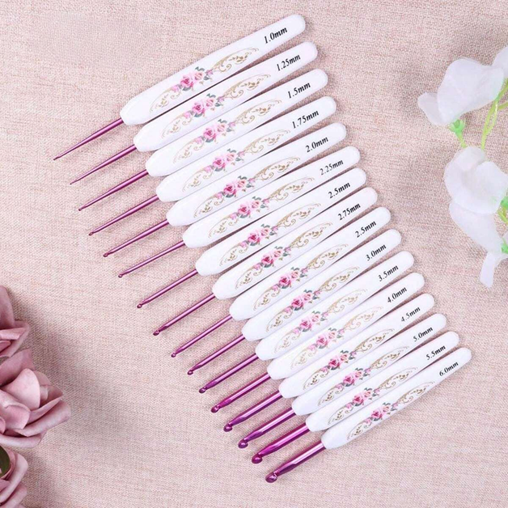 The Crochet Hook Set 16 Pcs with Case includes hooks with artistic white floral handles and pink metal tips, each size labeled and arranged diagonally on a light fabric background.