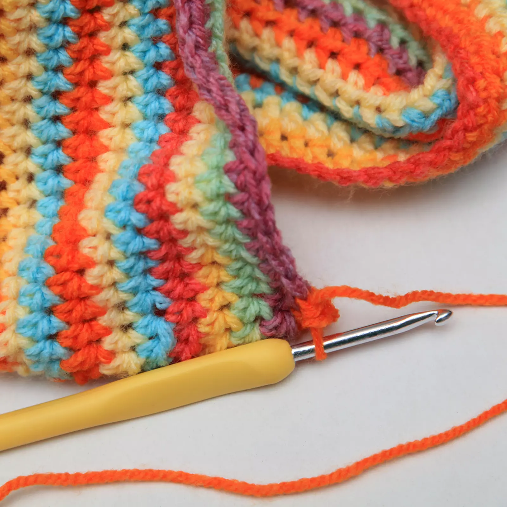 An Ergonomic Crochet Hook Set 9 Pcs, with a durable aluminum hook and yellow handle, rests on a vibrant striped crochet project featuring orange, yellow, pink, blue, and green yarn.