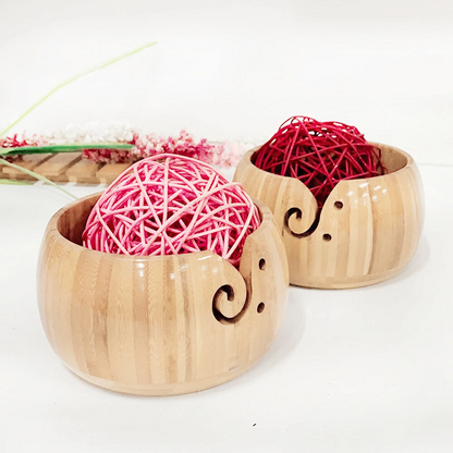 Two Wooden Yarn Bowls With Lid, each with carved swirl notches and a high gloss finish, hold pink and red yarn balls. Set on a white surface with dried flowers, they’re ideal for knitting projects.