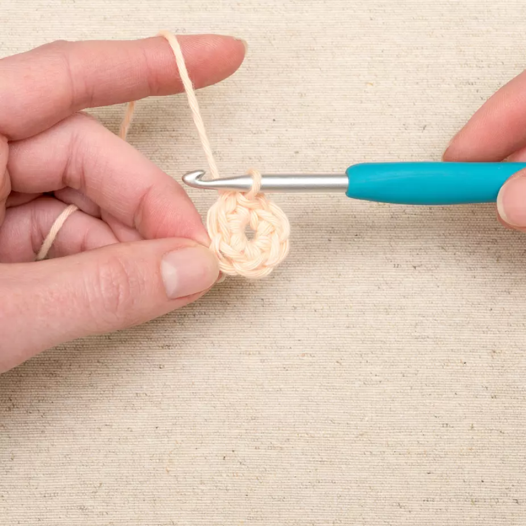 A person crochets a small beige circle using a soft-grip handle from the Ergonomic Crochet Hook Set 9 Pcs, with a beige fabric background.