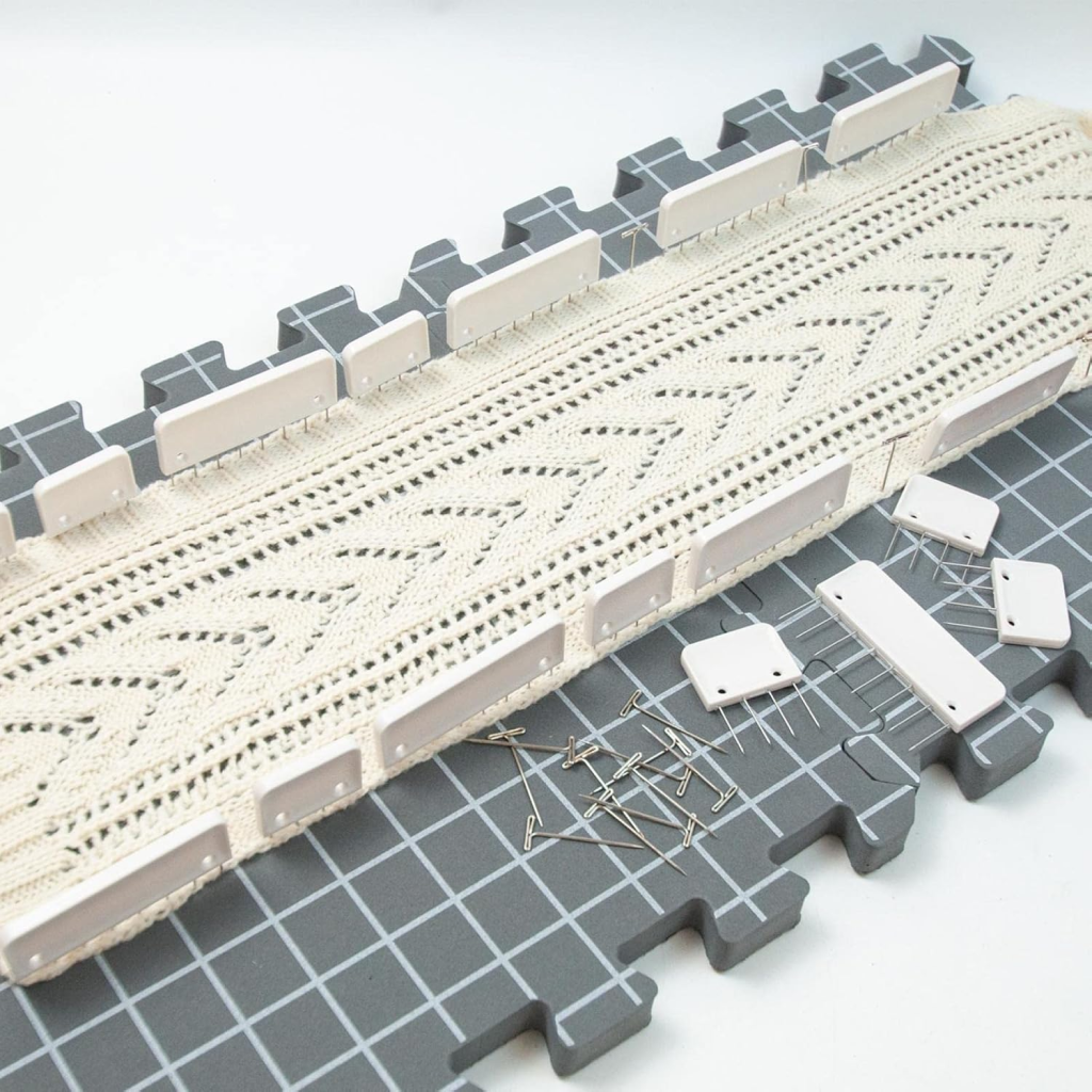 A lace strip is pinned to a gray blocking mat with the Knit Blocking Comb Set, showcasing precise blocking on white grid lines, while durable T-pins and extra combs from the set are scattered nearby.