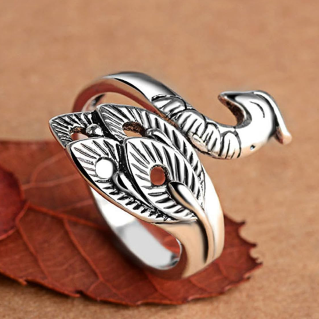 The Yarn Tension Control Ring, crafted from stainless steel and designed with leaf-shaped patterns that mimic a curled snake, is showcased on a brown surface with a dried leaf beneath it.