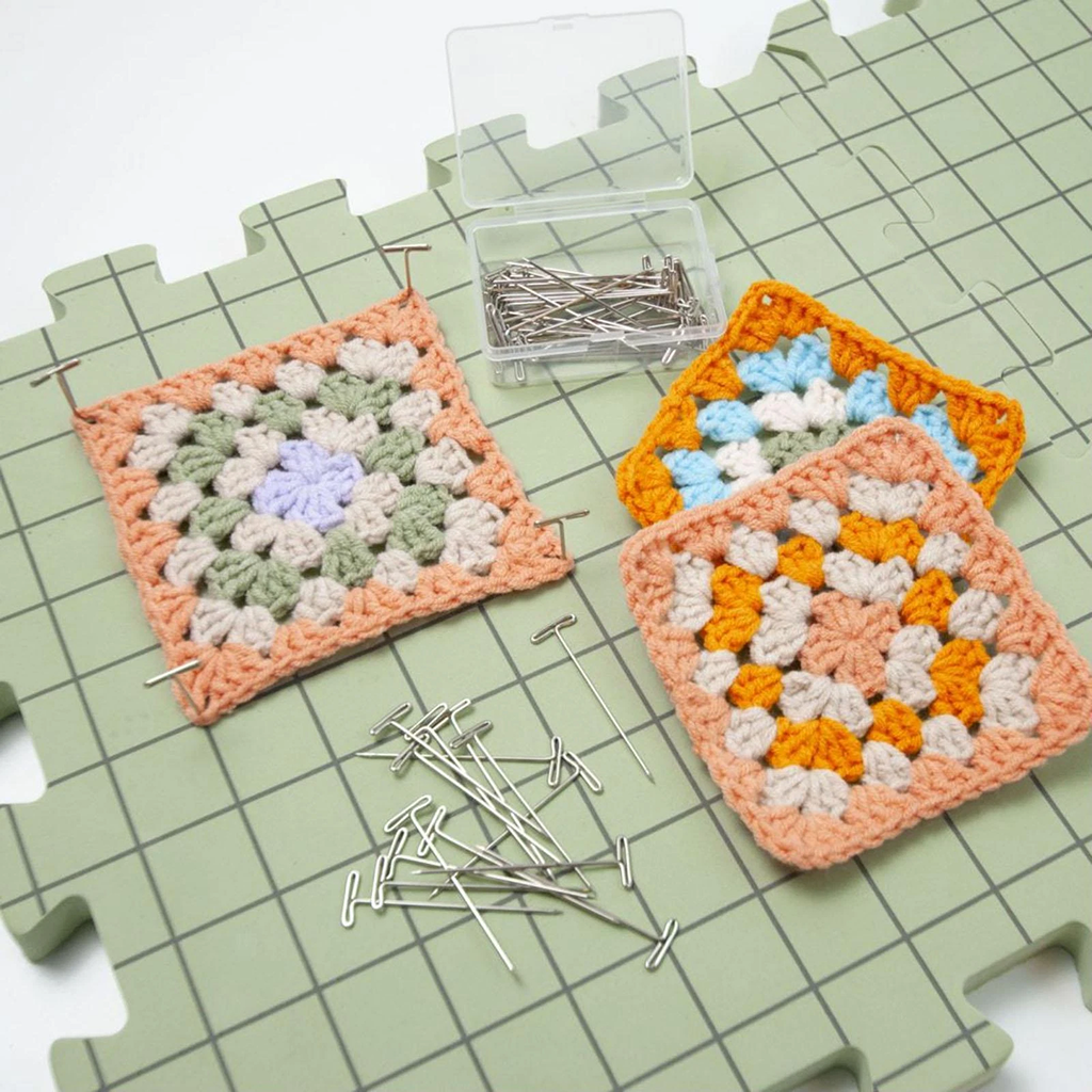 Three crocheted granny squares are pinned to Knitting Crocheting Blocking Mats with a green grid pattern, accompanied by a box of T-pins and some loose pins nearby.